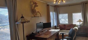 Une pièce comprenant un bureau en bois, un ordinateur, une chaise, un canapé, un élément décoratif représentant un cerf accroché au mur, et une fenêtre avec des rideaux.