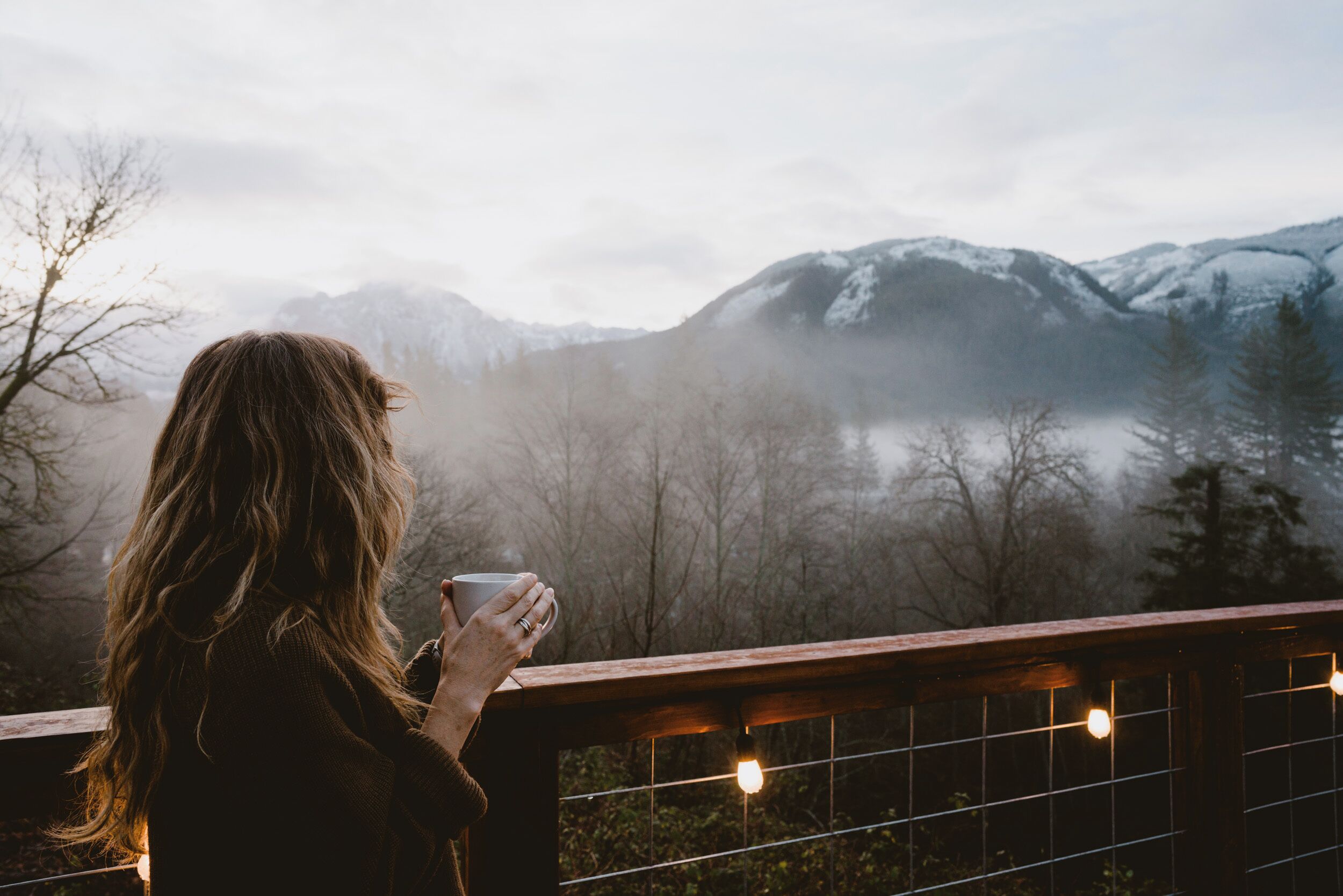 Una persona sosteniendo una taza, con vista a un paisaje montañoso con baranda de madera y luces colgantes.
