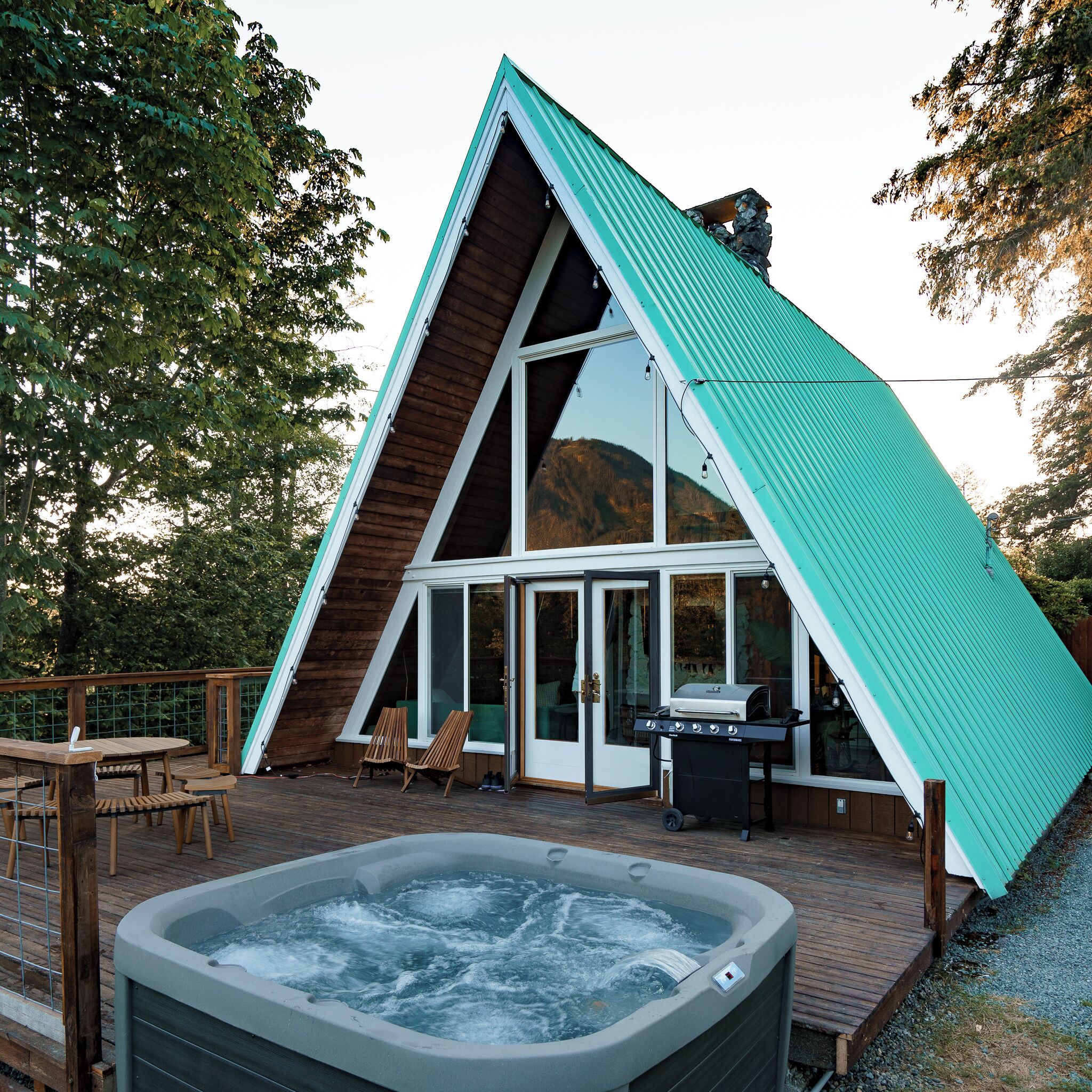 A modern A-frame building with a hot tub, deck chairs, and a grill area.