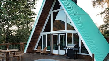 A modern A-frame building with a hot tub, deck chairs, and a grill area.