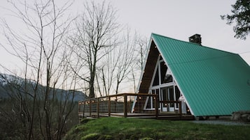 Una cabana moderna amb teulada triangular, terrassa de fusta i finestres de vidre, envoltada d'un paisatge forestal.