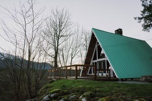 A modern, triangular-roofed cabin with a wooden deck and glass windows, surrounded by a forested landscape.