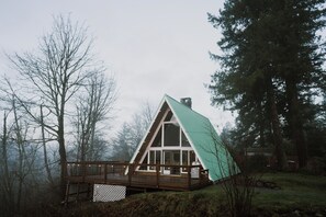 A cabin with a steeply pitched roof, large windows, and a wooden deck. - PNW A-Frame - Hot tub with view and A/C (Gold Bar)