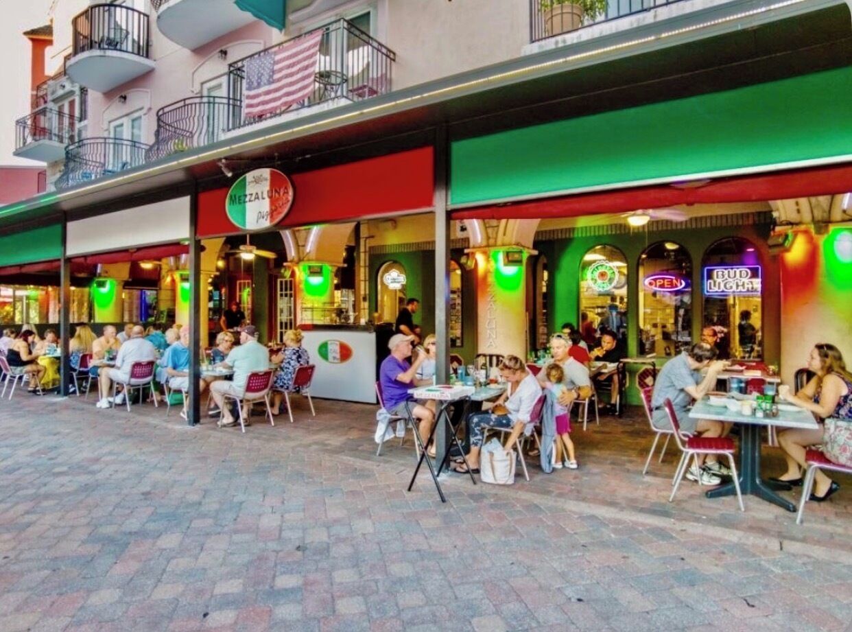 Un espace de restauration en plein air avec plusieurs tables et chaises, où l’on peut voir diverses personnes prendre leur repas, et une devanture de magasin avec des enseignes illuminées.
