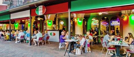 Un espace de restauration en plein air avec plusieurs tables et chaises, où l’on peut voir diverses personnes prendre leur repas, et une devanture de magasin avec des enseignes illuminées.