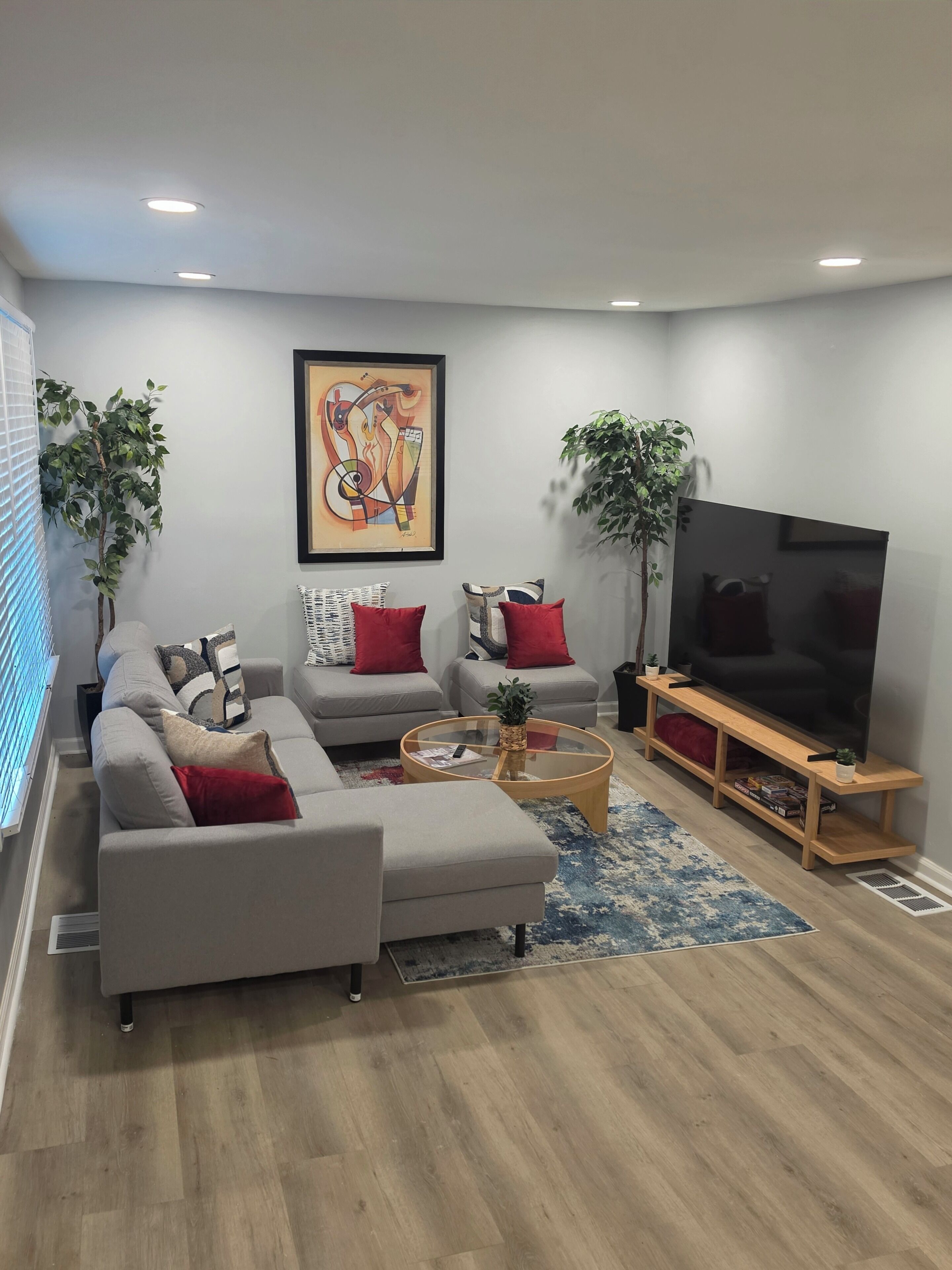 A modern living room with a grey sofa set, red cushions, a glass coffee table, and a large flat-screen TV on a wooden stand.