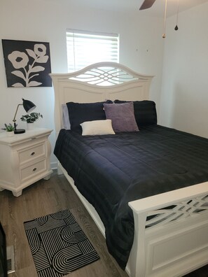 Quarto com cama, mesa de cabeceira, abajur e janela com persianas.