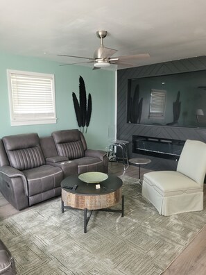 Uma sala de estar com sofá seccional, mesa de centro redonda, poltrona branca, lareira e ventilador de teto.
