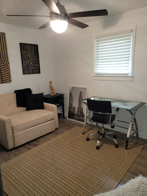 A room with a ceiling fan, a desk with a chair, a sofa, a rug, and artwork on the walls.