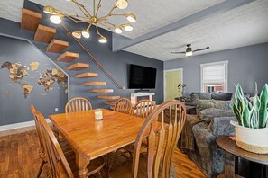 A dining area with a wooden table and chairs, a television mounted on the wall, and a staircase with wooden steps. - Luxury Modern 30+ Day Bungalow. Walk Downtown. Hot Tub. Pet Friendly (Yellow Springs)