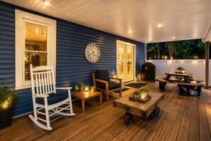 A covered outdoor patio with wooden flooring, a rocking chair, a table with chairs, and a barbecue grill. - Luxury Modern 30+ Day Bungalow. Walk Downtown. Hot Tub. Pet Friendly (Yellow Springs)
