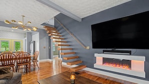 A modern living room with a wooden staircase, a fireplace, a flat-screen TV, and a dining area with wooden chairs. - Luxury Modern 30+ Day Bungalow. Walk Downtown. Hot Tub. Pet Friendly (Yellow Springs)