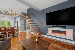 A modern living room with a wooden staircase, a fireplace, a flat-screen TV, and a dining area with wooden chairs. - Luxury Modern 30+ Day Bungalow. Walk Downtown. Hot Tub. Pet Friendly (Yellow Springs)