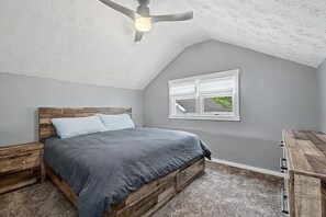 A bedroom with a sloped ceiling, a bed with a wooden headboard, a window with a view, and a ceiling fan. - Luxury Modern 30+ Day Bungalow. Walk Downtown. Hot Tub. Pet Friendly (Yellow Springs)