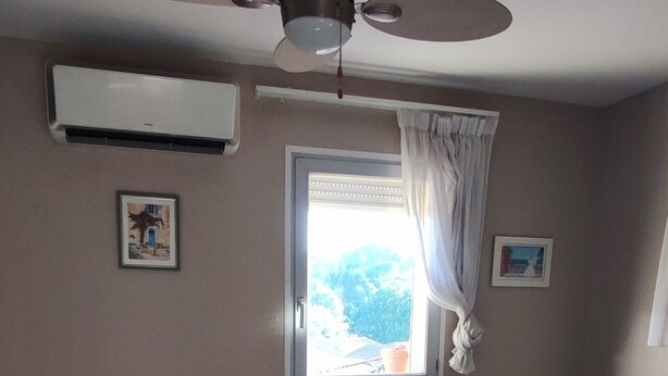 A bedroom with a bed, a ceiling fan, an air conditioning unit, a window with curtains, and a nightstand with a lamp. - T2 climatisé avec vue proche de Cannes et Grasse (Mouans-Sartoux)
