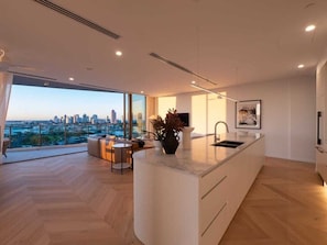 A modern kitchen with a large island, a dining area with a view, and a cityscape visible through large windows. - The Arches by Aryve | Surfers Paradise (Surfers Paradise)
