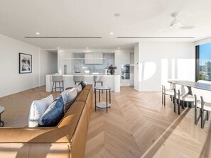 A modern living space with a herringbone floor pattern, a sofa with cushions, a dining area with a table and chairs, and a kitchen area with white cabinetry and a central island. - The Arches by Aryve | Surfers Paradise (Surfers Paradise)