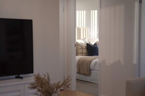 A room with a television on a stand, a bed with a blanket and pillows, and a window with blinds. - Sundays by Aryve | Ocean Terrace, Palm Beach (Palm Beach)