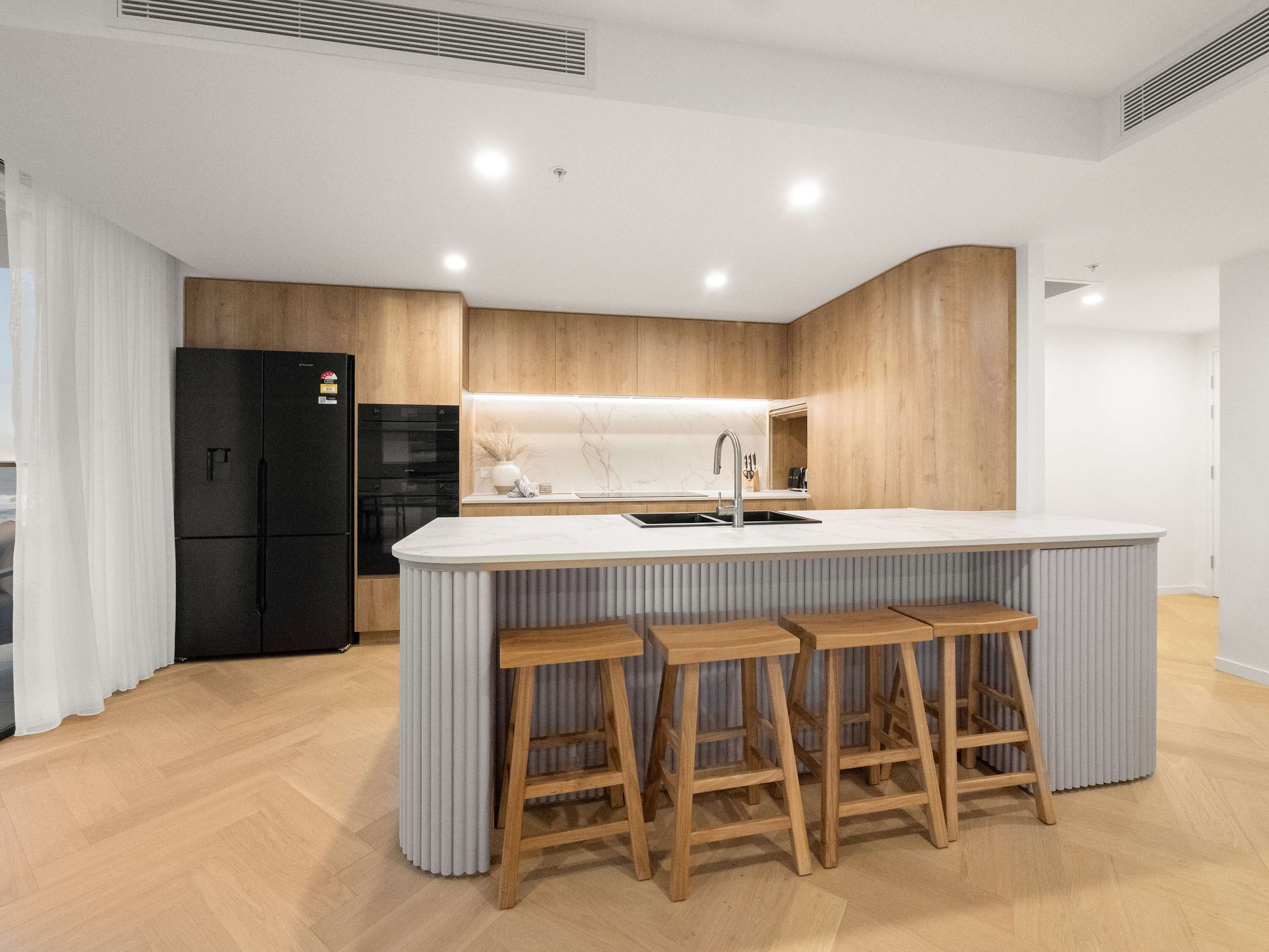 Una cocina moderna con una isla central, taburetes de madera, un refrigerador negro y una encimera blanca.