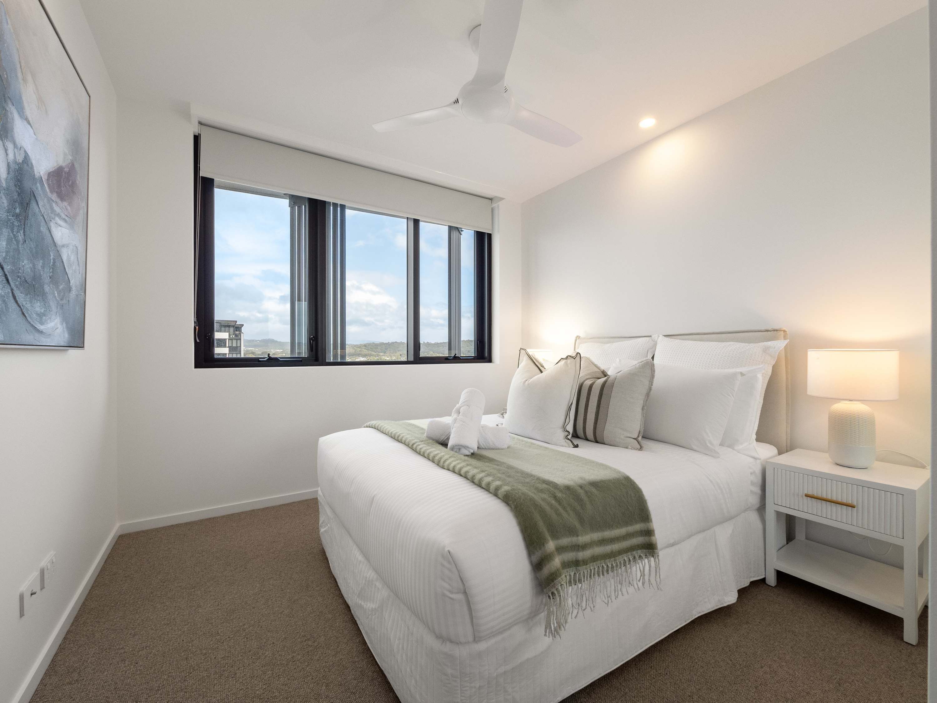 Un dormitorio con cama, mesita de noche, lámpara, ventana y ventilador de techo.