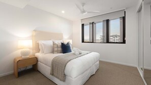 Quarto moderno com cama, mesa de cabeceira, abajur e vista para edifícios pela janela.