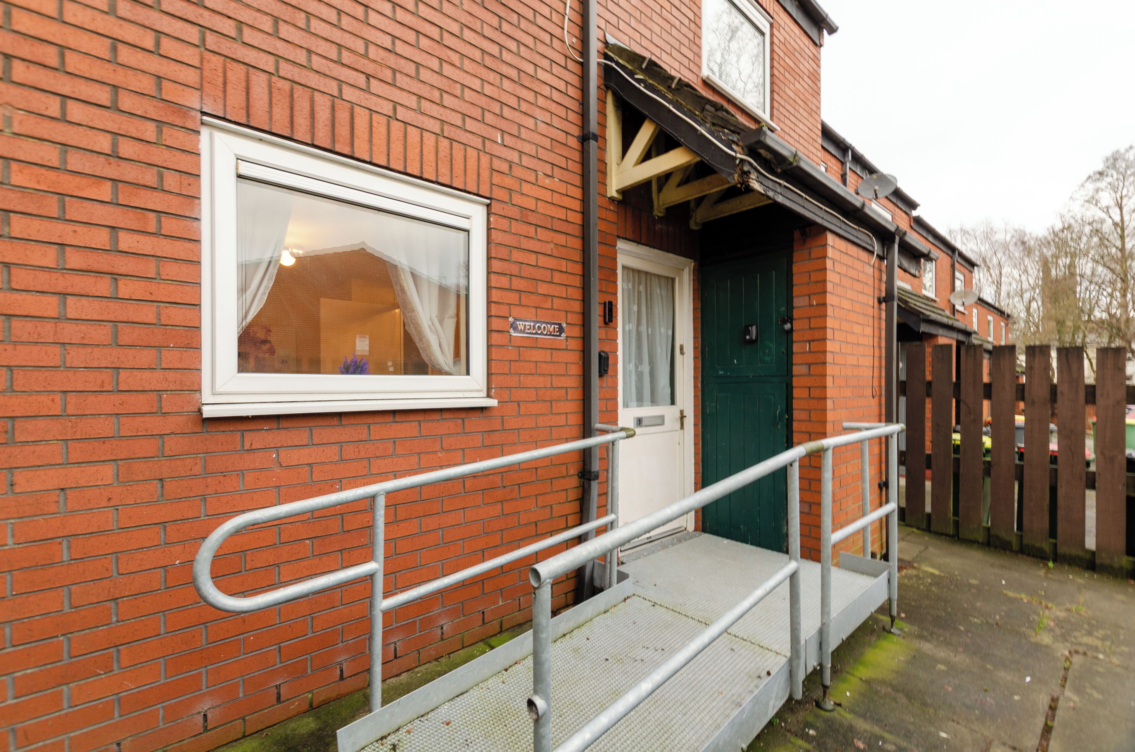 A red brick building with a white door, a 'Welcome' sign, and a ramp leading to the entrance.