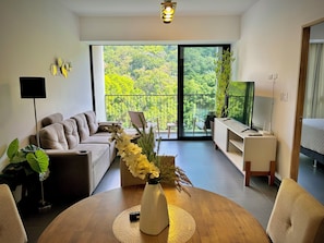 Ein modernes Wohnzimmer mit einer Couch, einem Couchtisch mit Blumenvase, einem Fernseher und einem Balkon mit Blick auf Grünflächen.