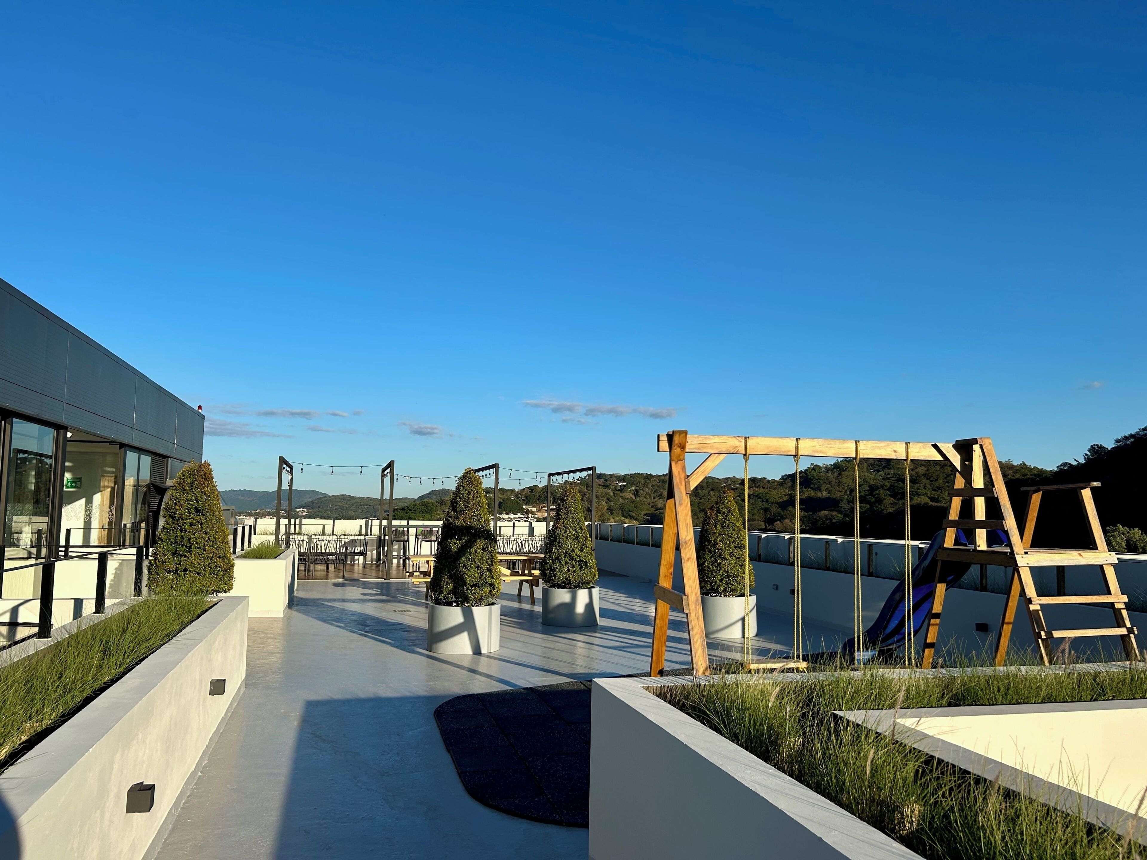 Una terraza en la azotea con un edificio moderno, plantas en macetas y un conjunto de columpios de madera.