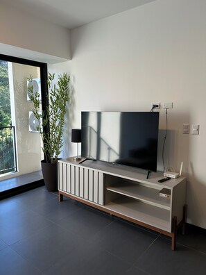 Ein modernes Wohnzimmer mit einem Flachbildfernseher auf einer schlichten Ablage, einer Zimmerpflanze und einem großen Fenster mit Blick auf Grünflächen.