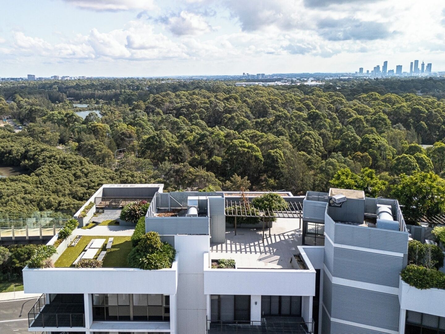 A modern building with a rooftop garden, surrounded by dense forest and a distant city skyline.