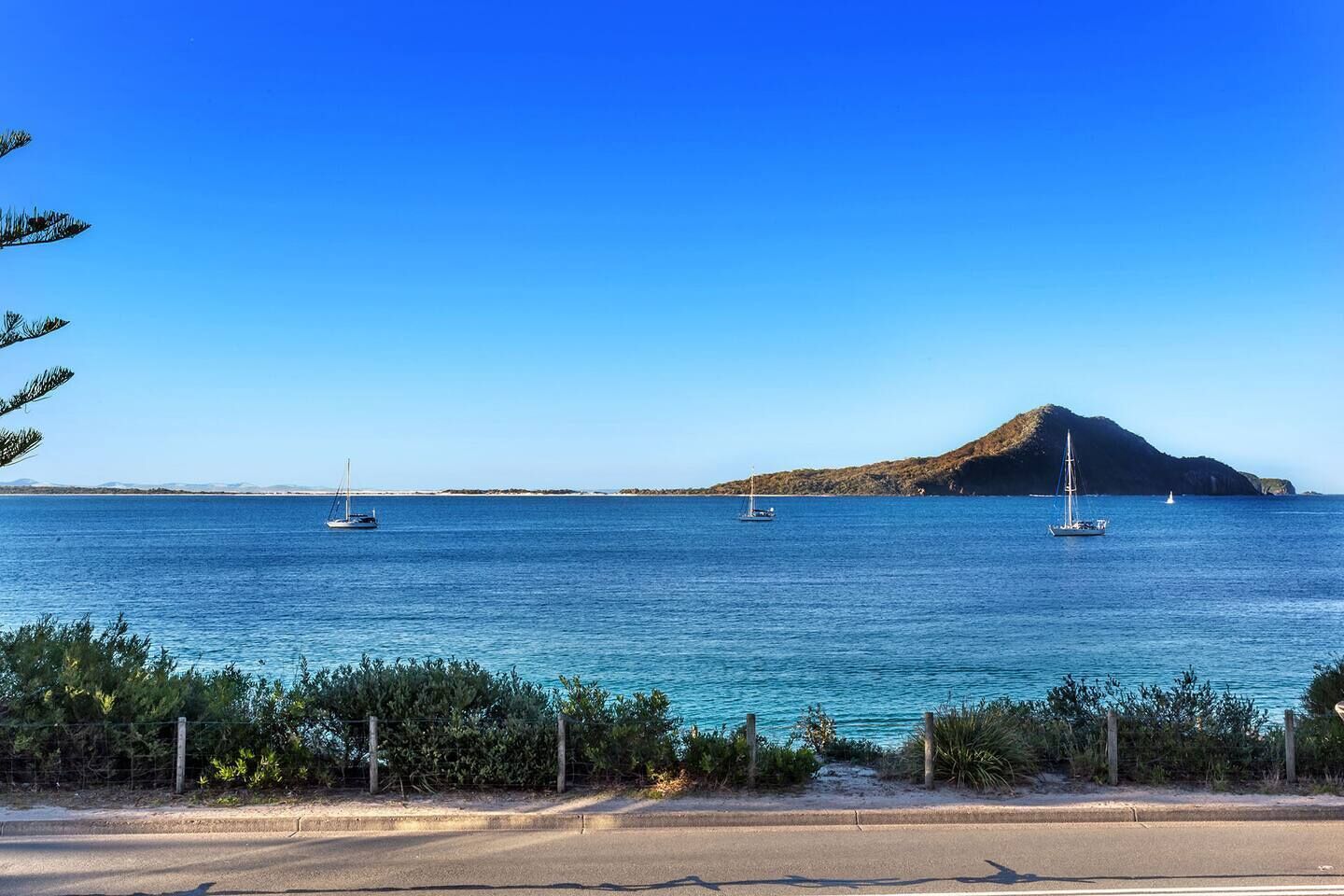 A coastal road with a clear view of the sea, three sailboats, and a prominent hill in the background.