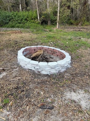 Ein runder Feuerkorb mit Ziegelbordurand, umgeben von trockenen Blättern und Zweigen.