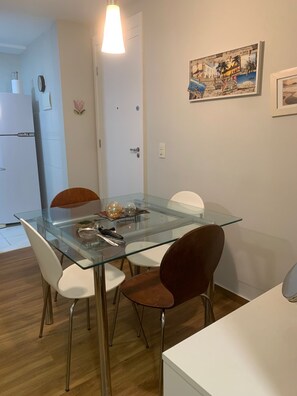 A dining area with a glass-top table, white chairs, and a white cabinet. - Olympic Bar well located (Rio de Janeiro)