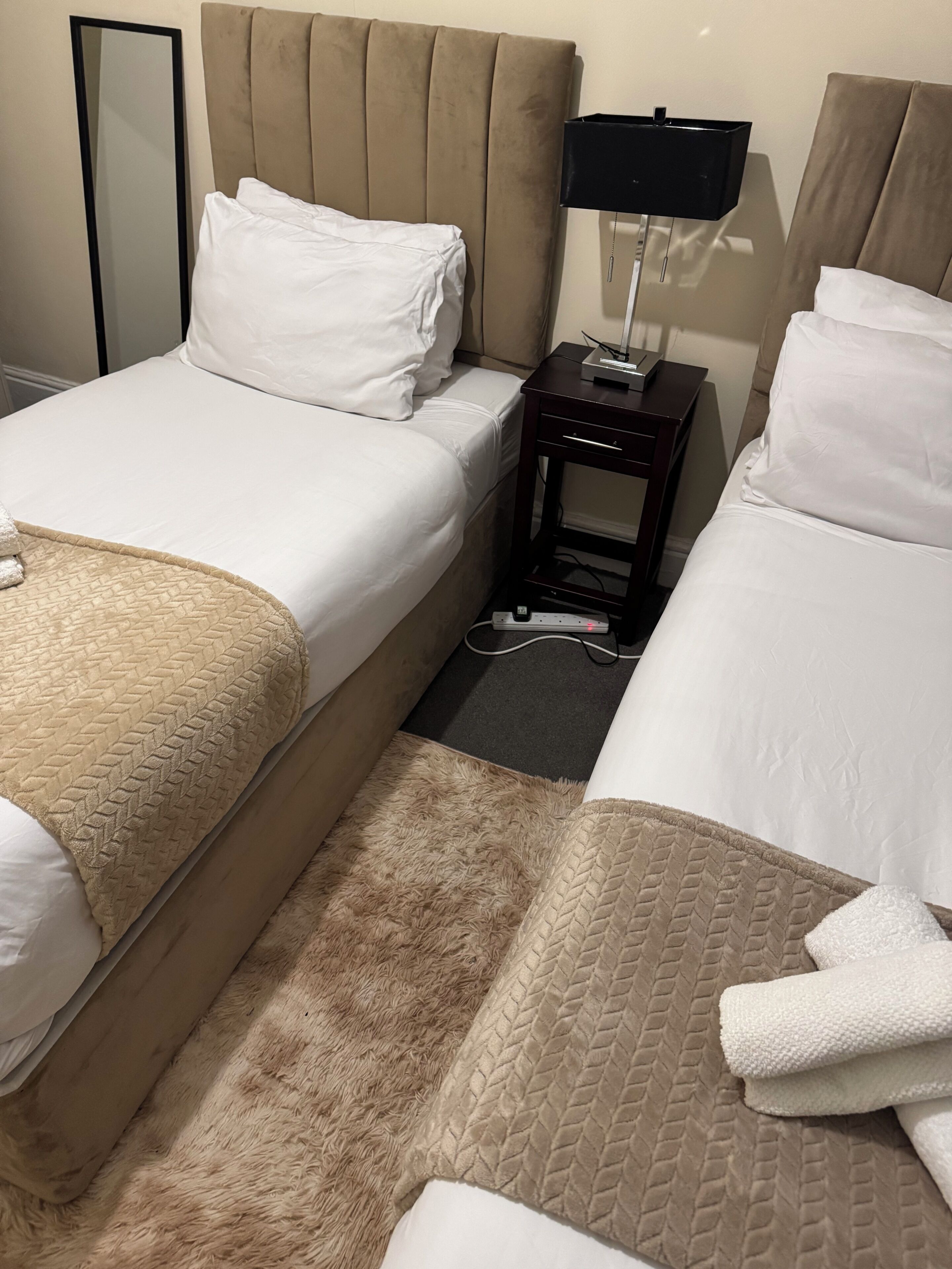 A hotel room with two beds, a bedside table with a lamp, and a mirror.