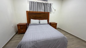 A bedroom with a wooden headboard, a grey bedspread, two bedside tables, and a grey curtain.