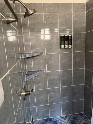 Ein Duschbereich mit gefliesten Wänden, einer Handbrause, einem am Wand montierten Wasserhahn und einer Ablage mit Toilettenartikeln.