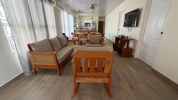 A living room with a sofa, chairs, a wooden bench, a dining table, and a kitchen in the background.