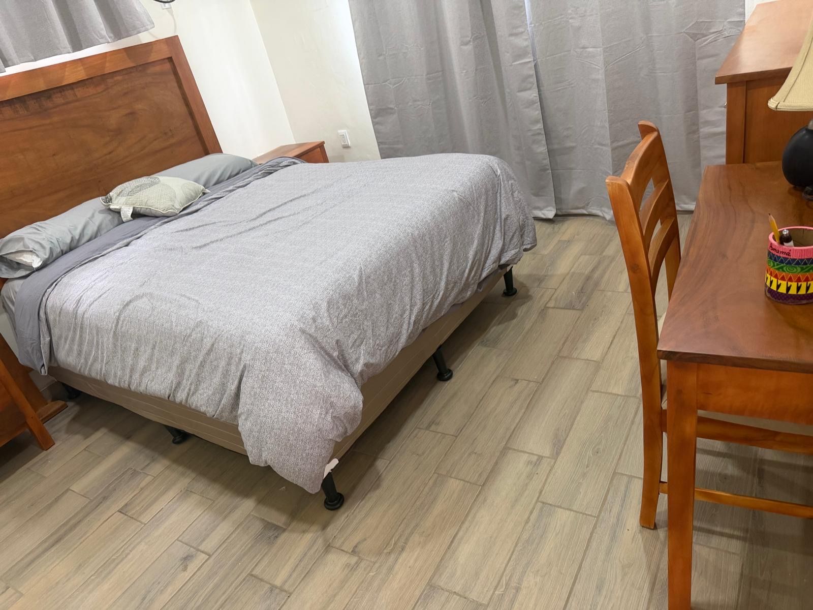 Un letto singolo con copripiumino grigio e cuscini, un comodino in legno, un tavolino con una tazza e un cucchiaino, e una sedia.
