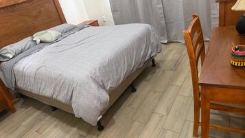 Una cama individual con colcha y almohadas grises, una mesita de noche de madera, una mesa de madera con una taza y una cuchara, y una silla.