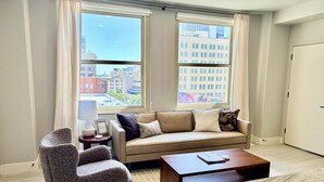 Una sala de estar moderna con sofá, sillón y mesa de centro, ventanas amplias con vistas a la ciudad y una puerta.
