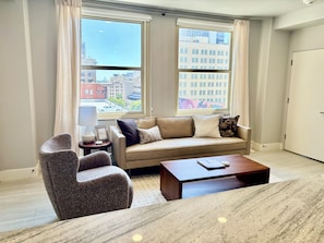 A modern living room with a sofa, armchair, and coffee table, large windows with city views, and a door.