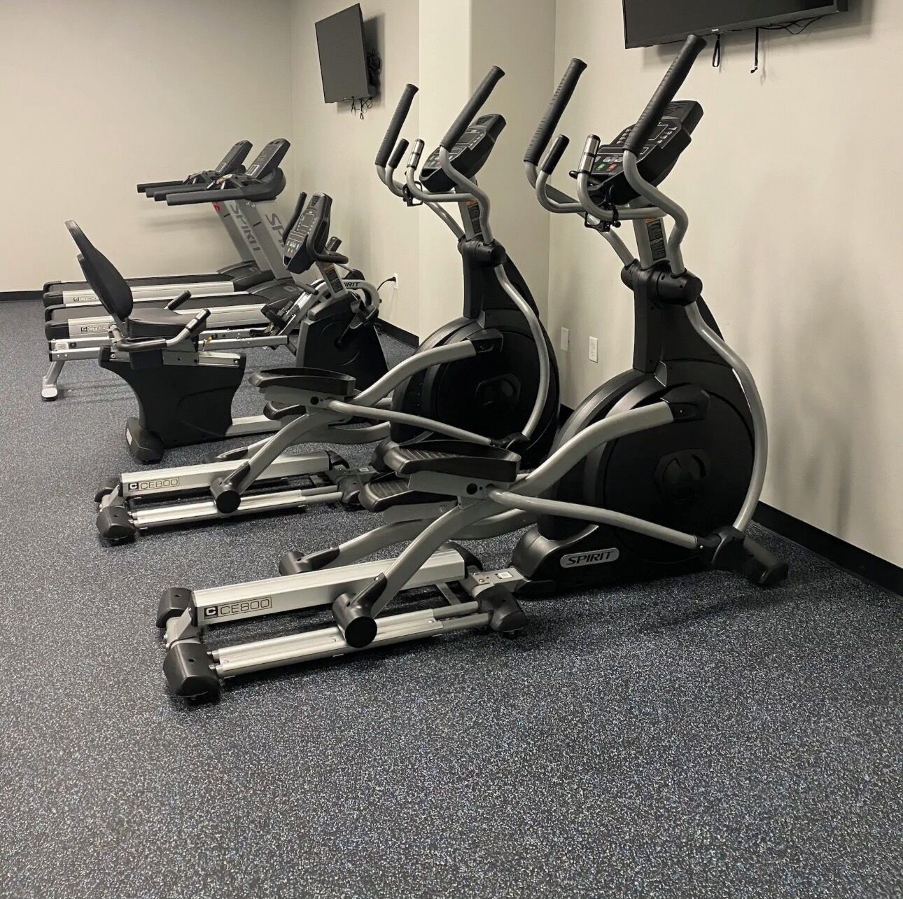 A row of stationary bikes in a gym with a carpeted floor and a plain wall.
