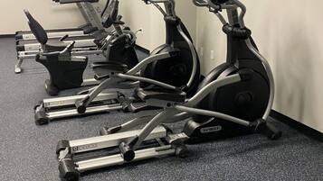 A row of stationary bikes in a gym with a carpeted floor and a plain wall.