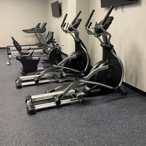 Een rij stilstaande fietsen in een sportschool met een vloerbedekking en een effen muur.