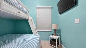 Una habitación con literas, una mesita con lámpara, un televisor montado en la pared y una ventana con persianas.