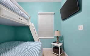 Ein Zimmer mit einem Etagenbett, einem kleinen Tisch mit Lampe, einem an der Wand montierten Fernseher und einem Fenster mit Jalousien.