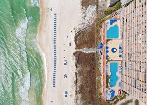Vue aérienne d’un hôtel situé en bord de mer, avec une longue rangée de parasols et de chaises longues le long du rivage, une piscine et un bâtiment doté d’une architecture originale.