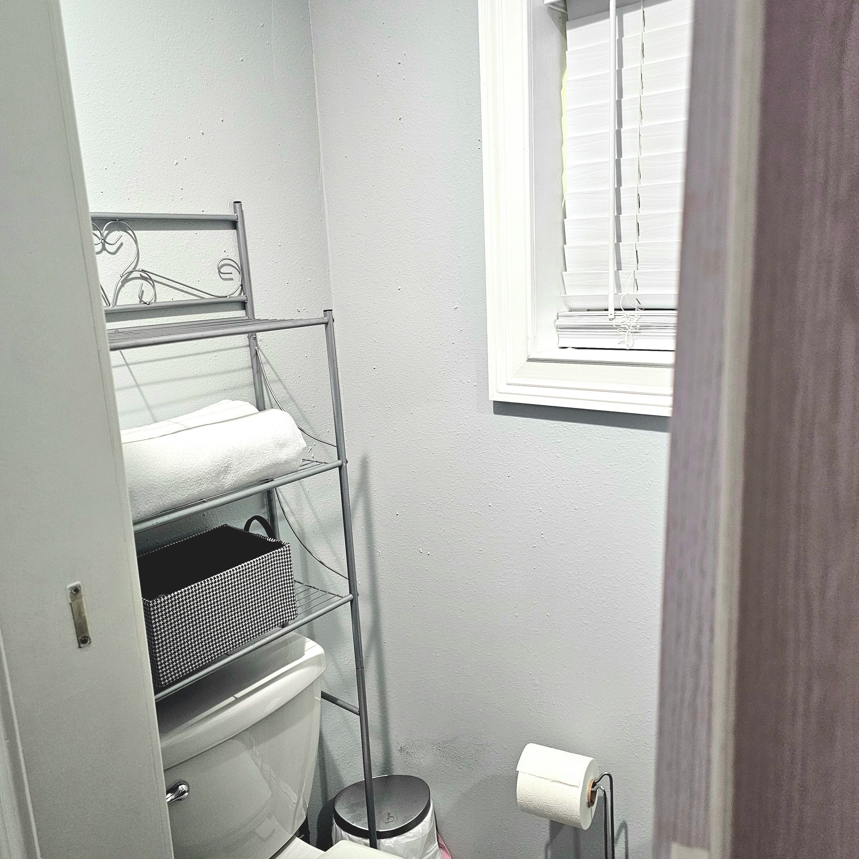 Ein Badezimmer mit einer weißen Toilette, einer Metallregaleinheit, einem Fenster mit Jalousien und einer Toilettenpapierrolle an der Wand.