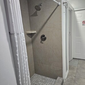 Un espace douche équipé d’un pommeau de douche à main, d’une étagère d’angle et d’une porte sur laquelle est apposé un panneau « Interdiction de fumer ».