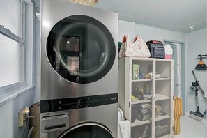 A compact, modern laundry room with a large, stainless steel washer and dryer, a storage cabinet with shelves, and a vacuum cleaner. - Cozy 3BR Sarasota Bungalow near Lido & Siesta (Sarasota)